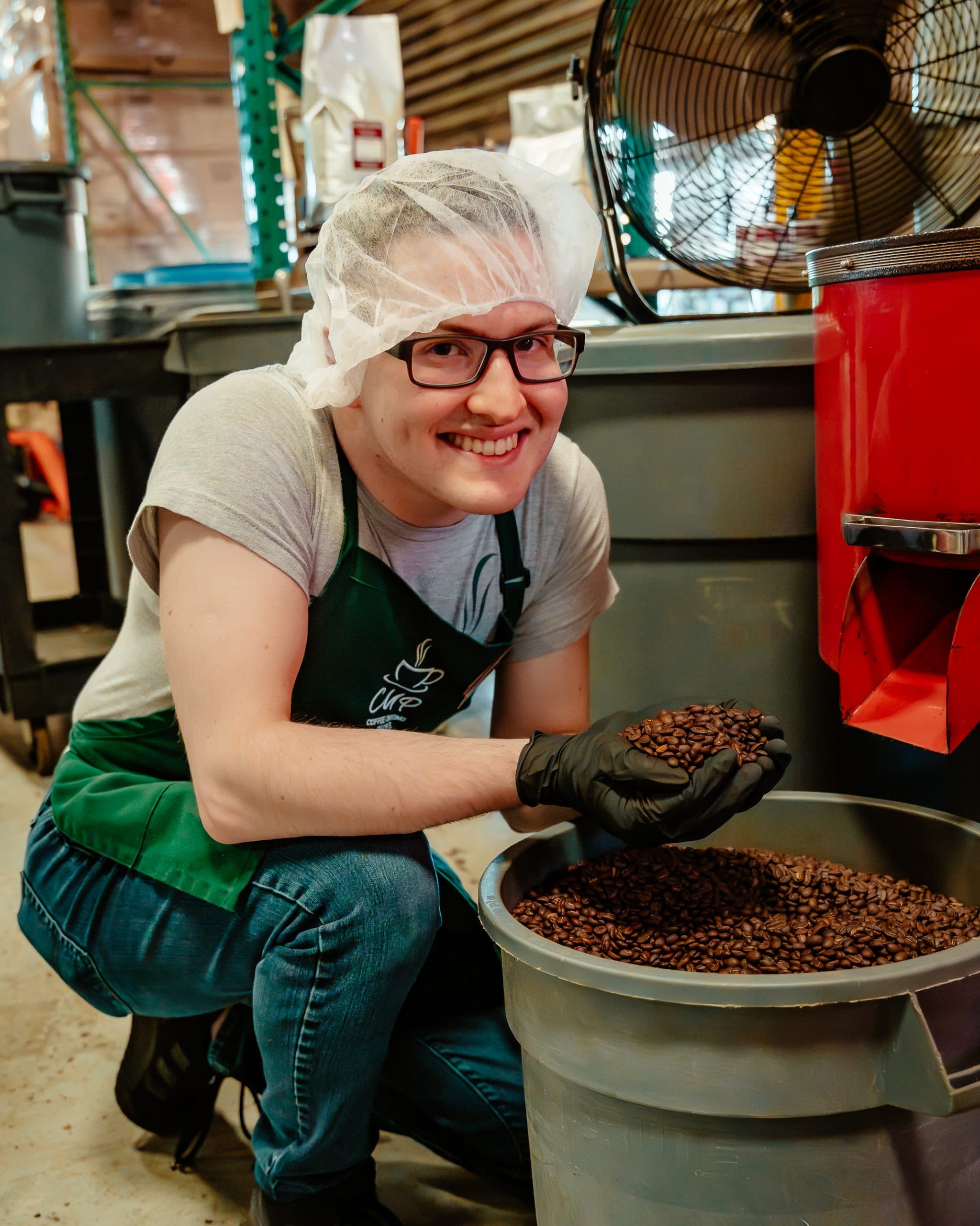 A CUP team member in a hairnet and apron scooping fresh beans from a roasting drum
